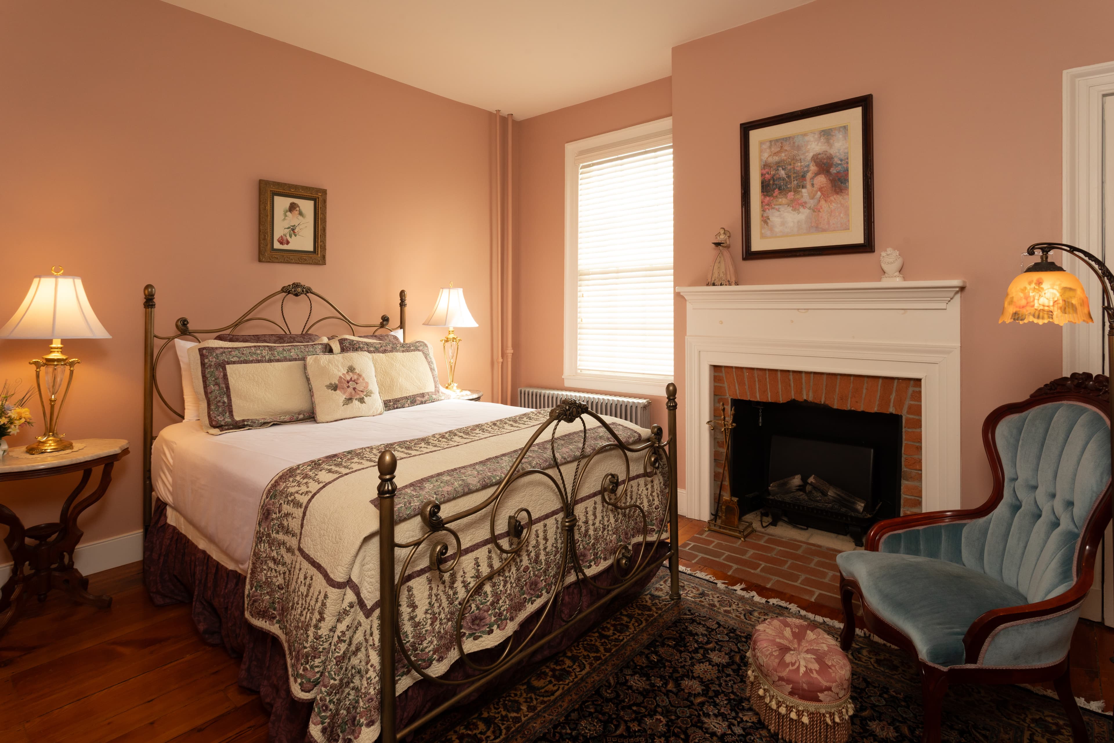 A cozy bedroom featuring a wrought iron bed, soft pink walls, a decorative fireplace, and elegant furnishings.