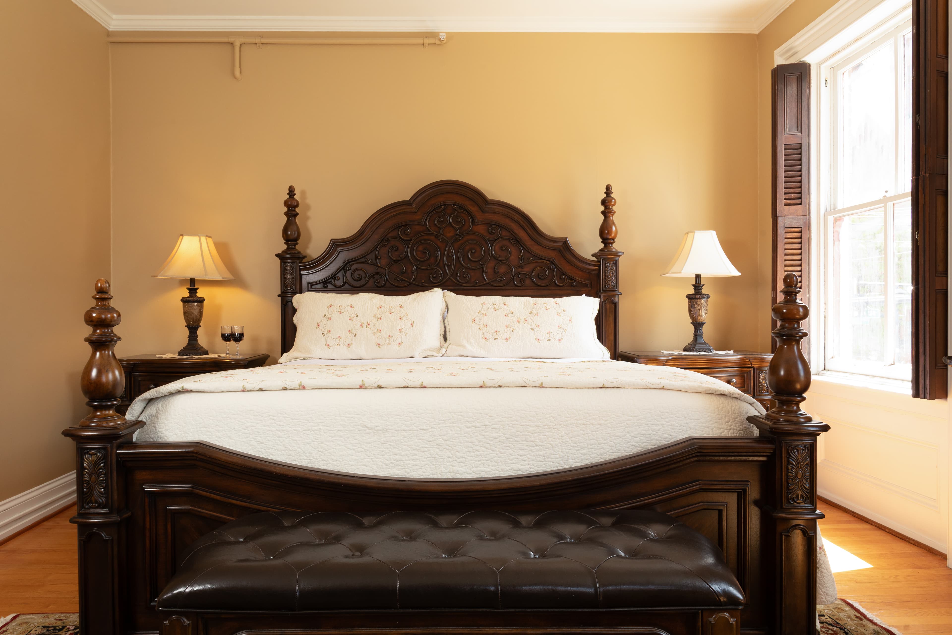 A beautifully decorated bedroom featuring a carved wooden bed, two lamps, and natural light from a window.