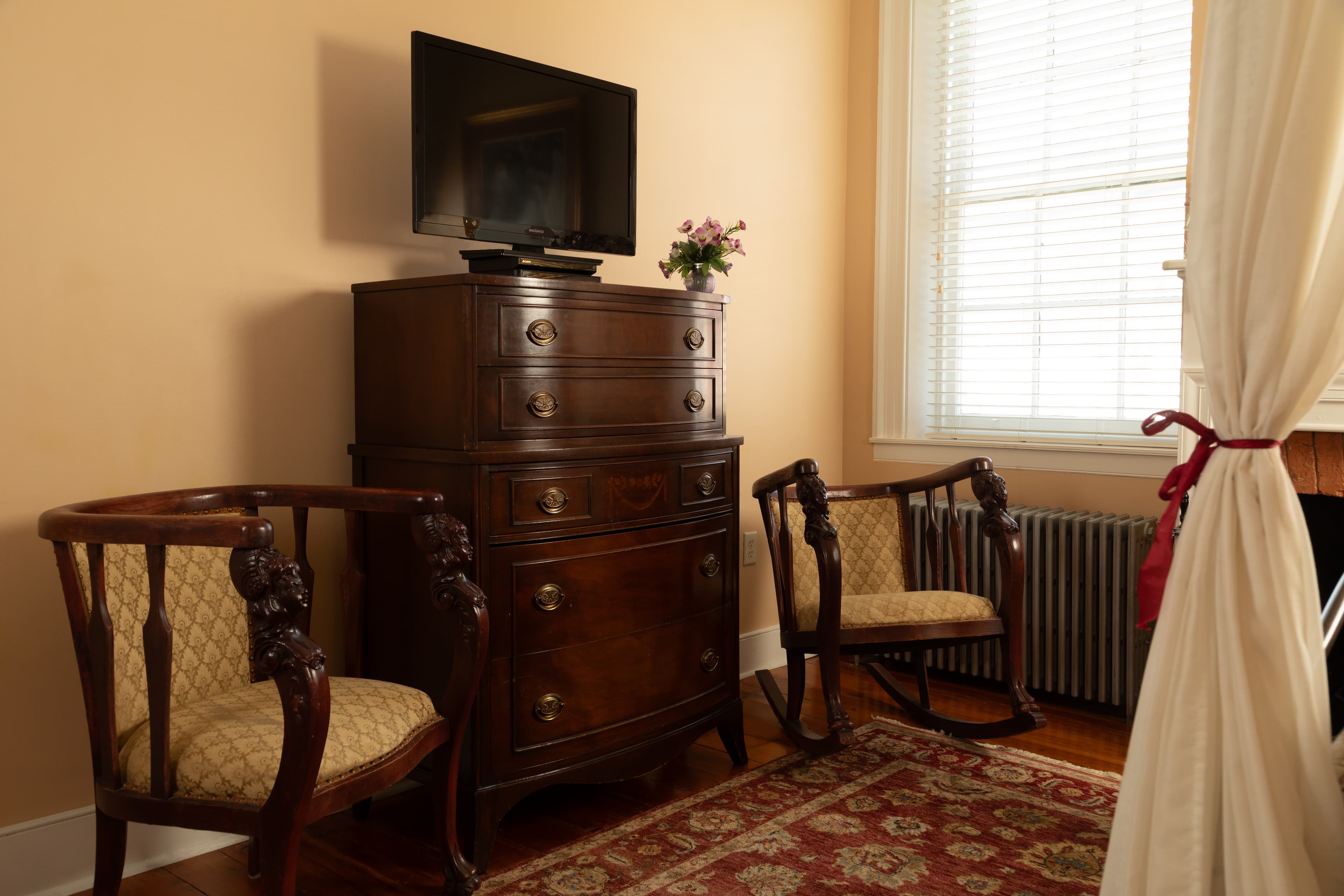 A cozy room featuring a wooden dresser with a TV on top, two elegant chairs, and a window with natural light.