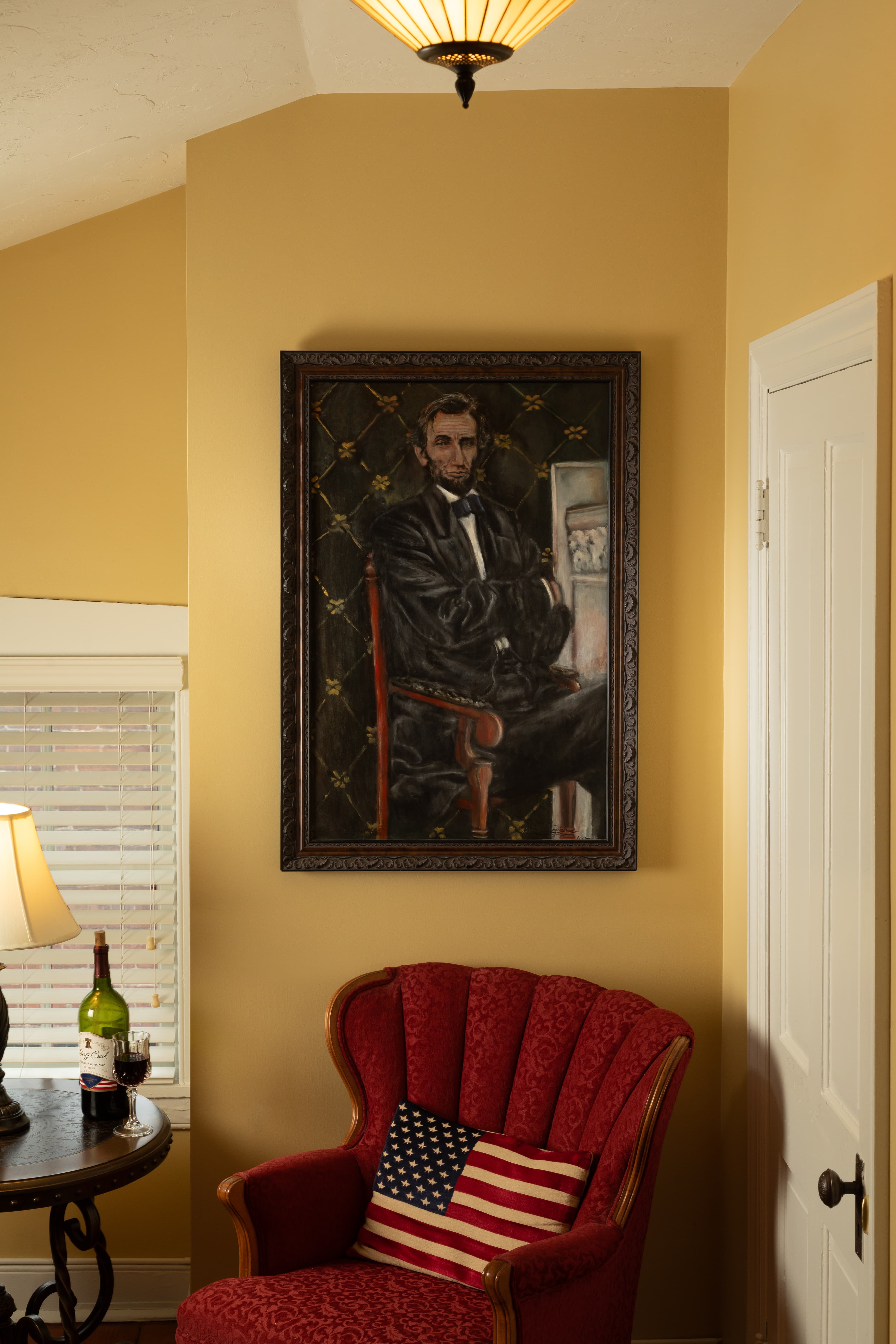 A cozy corner featuring a red upholstered chair, a side table with a lamp and wine, and a portrait of Abraham Lincoln on the wall.