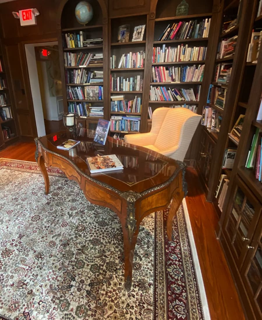 A cozy library featuring a wooden table, plush chair, and shelves filled with books.
