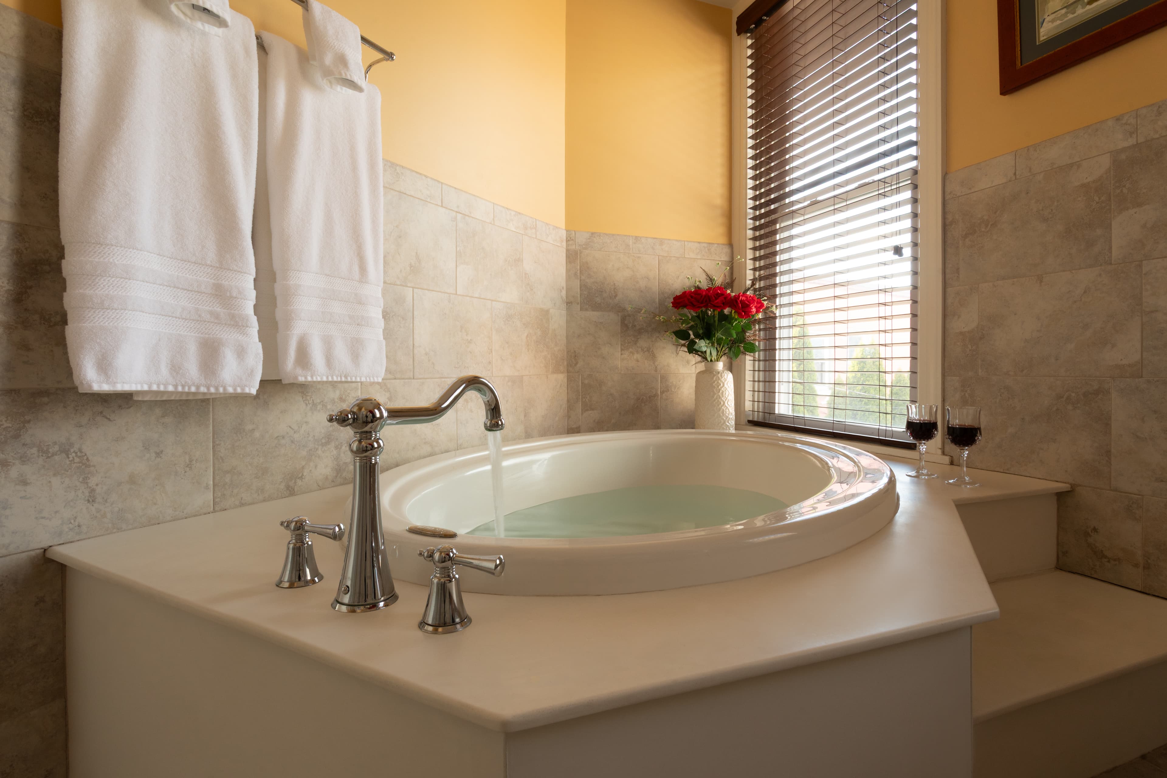 A luxurious bathtub with running water, surrounded by plush towels, a vase of roses, and two glasses of wine.