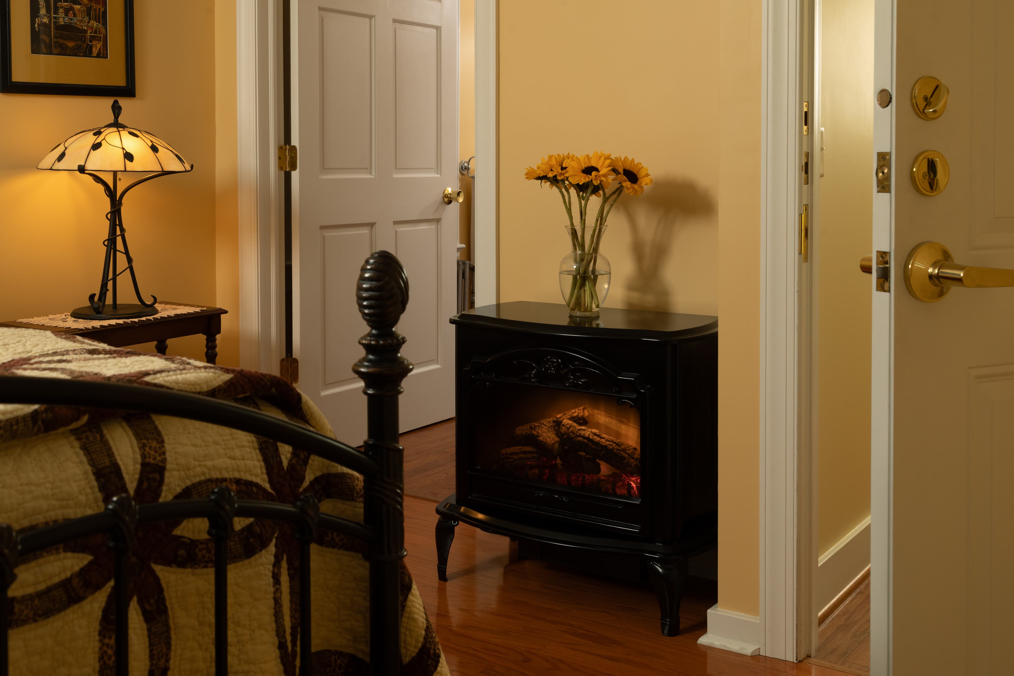 A cozy bedroom scene featuring a decorative lamp, a vase of sunflowers, and a black electric fireplace against a warm wall.