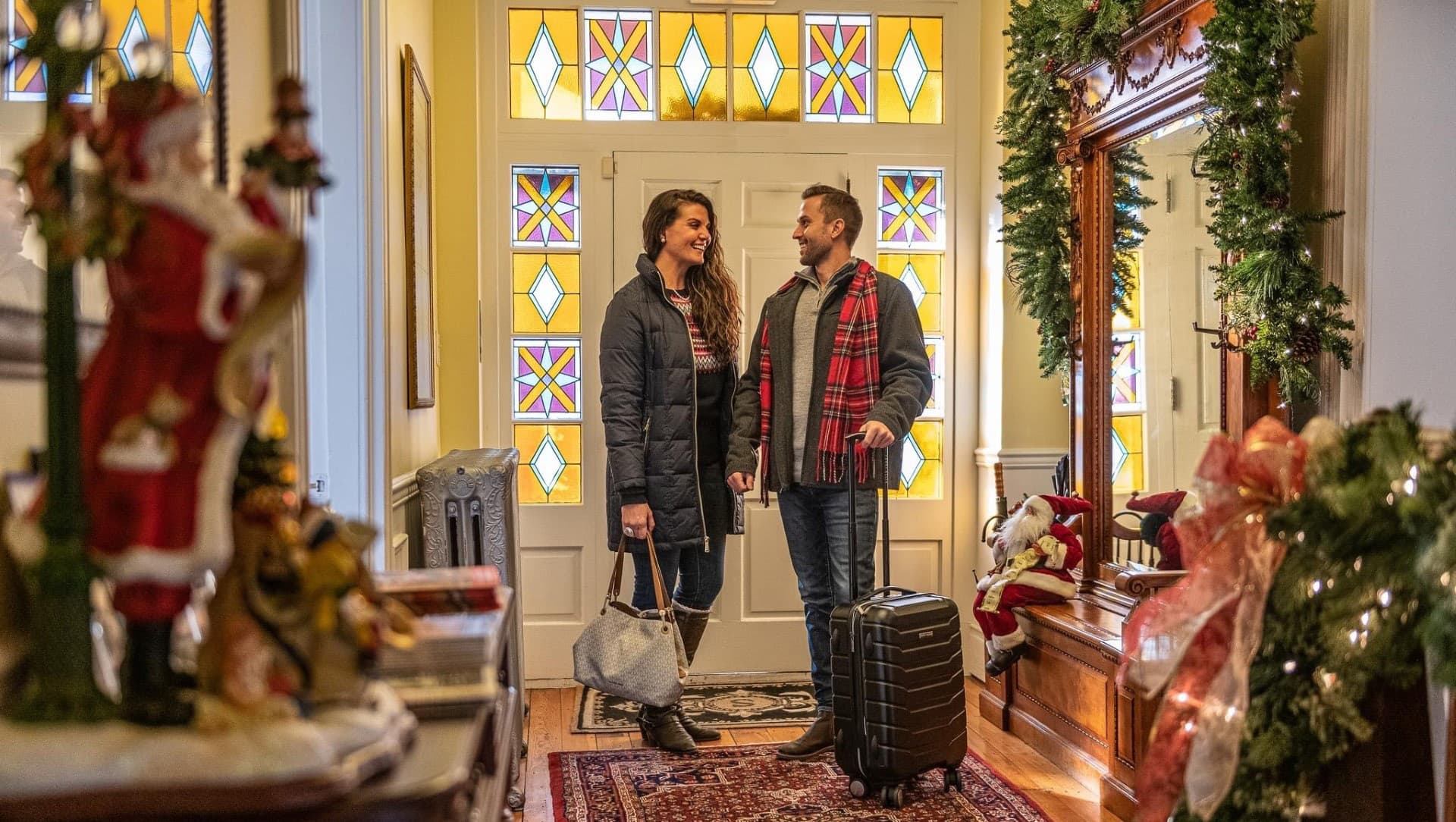 A smiling couple stands in a warmly decorated entrance hall with luggage, surrounded by holiday decor.
