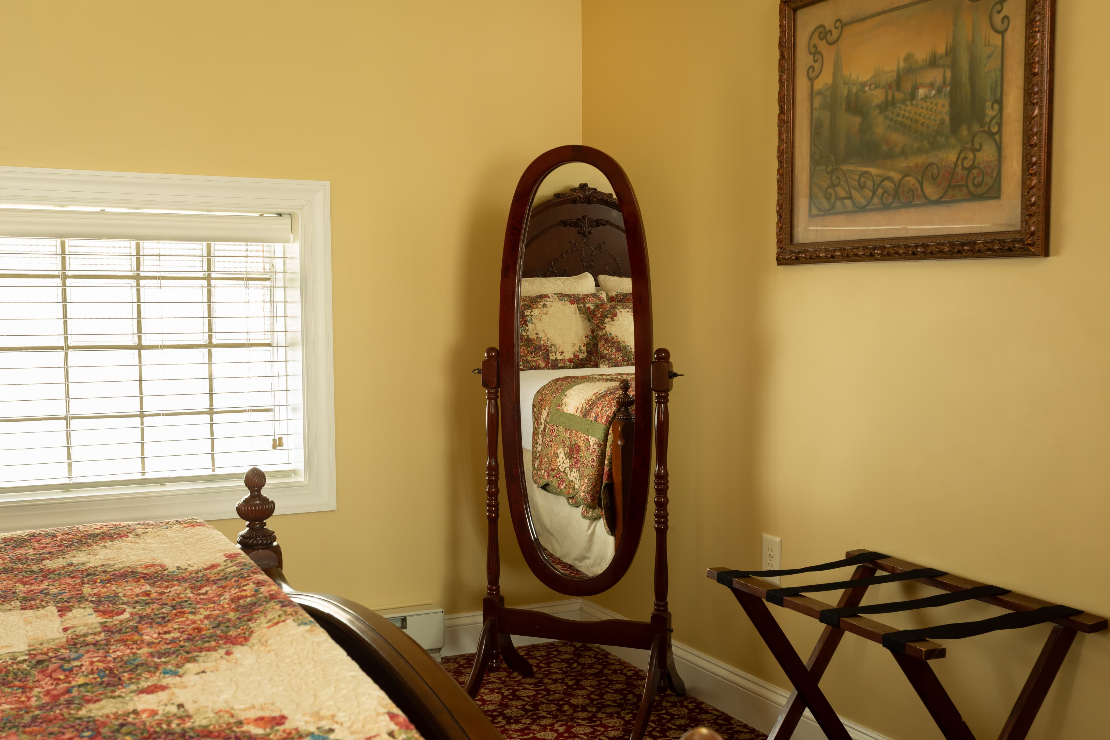 A cozy bedroom corner featuring a wooden mirror, a quilted bed, a small folding table, and a painting on the wall.