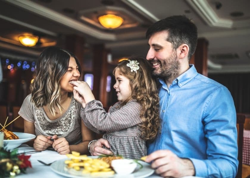A happy family enjoys a meal together in a restaurant, with a mother and father laughing as their daughter playfully feeds one of them. A happy family enjoys a meal together in a restaurant, with a mother and father laughing as their daughter playfully feeds one of them.