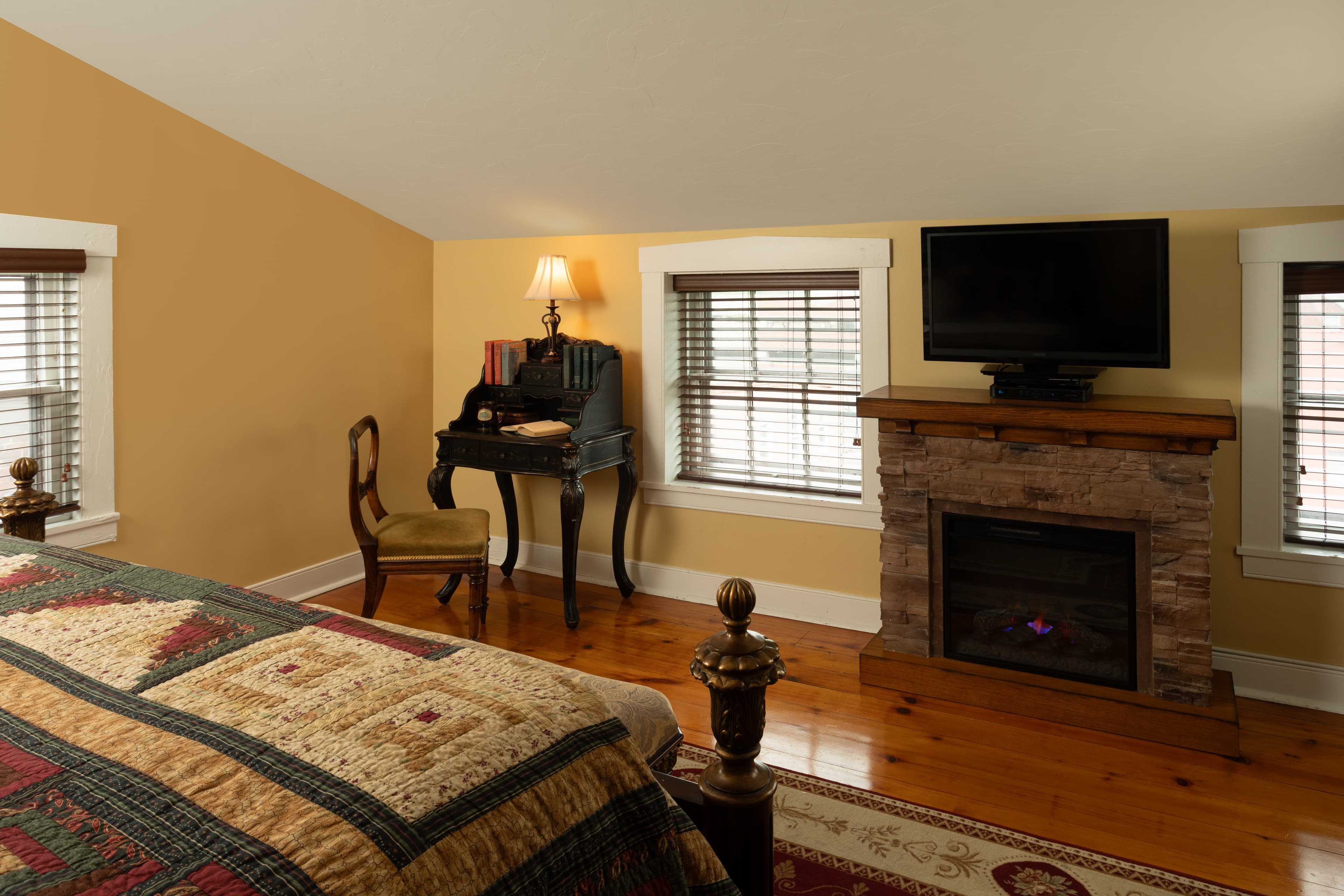 Cozy bedroom featuring a quilted bed, a fireplace, a TV, and a writing desk.