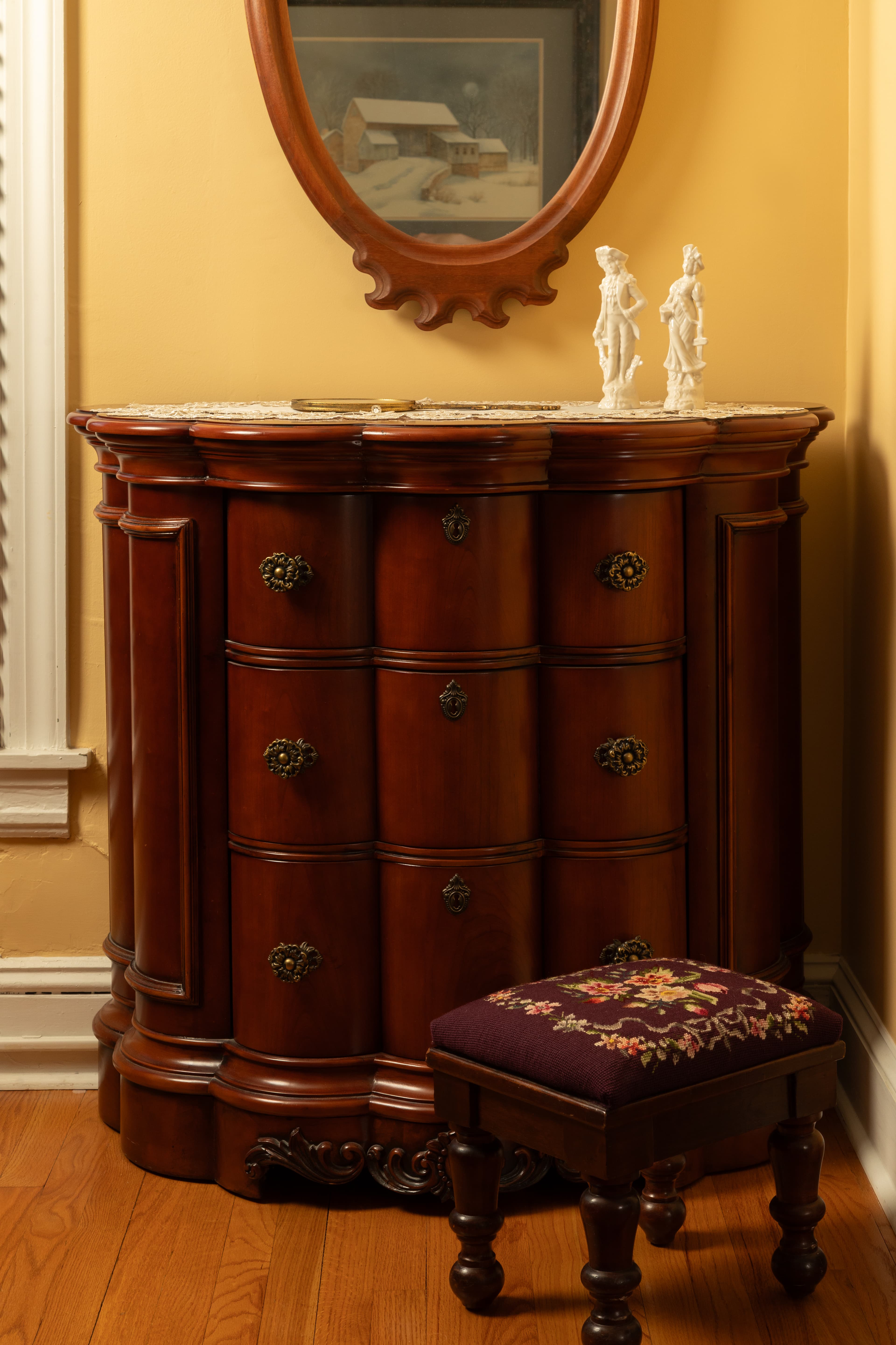 A wooden chest of drawers with ornate handles next to a stool, under a mirror, against a yellow wall.