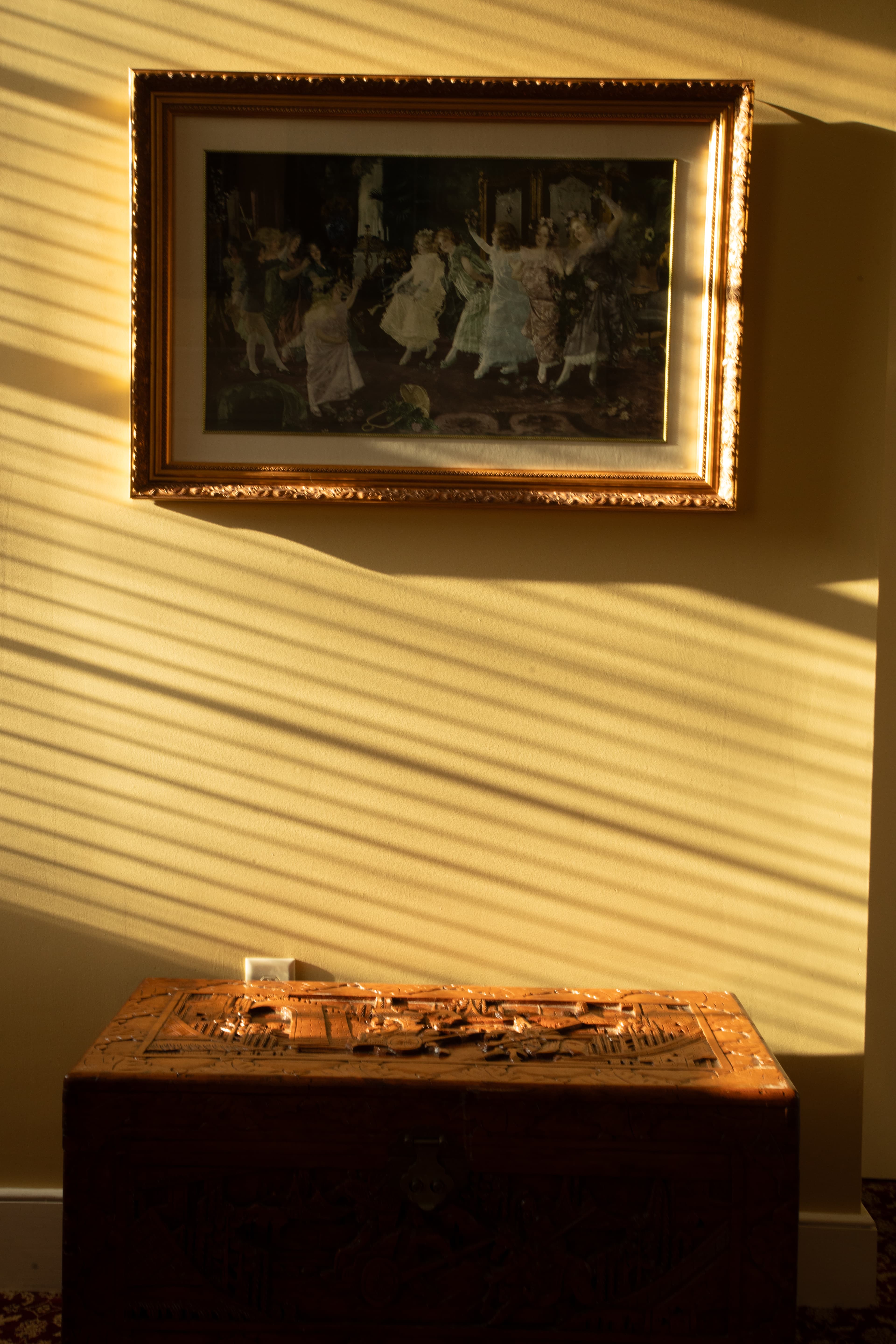 A vintage painting hangs above an ornate wooden chest, illuminated by streaks of sunlight.