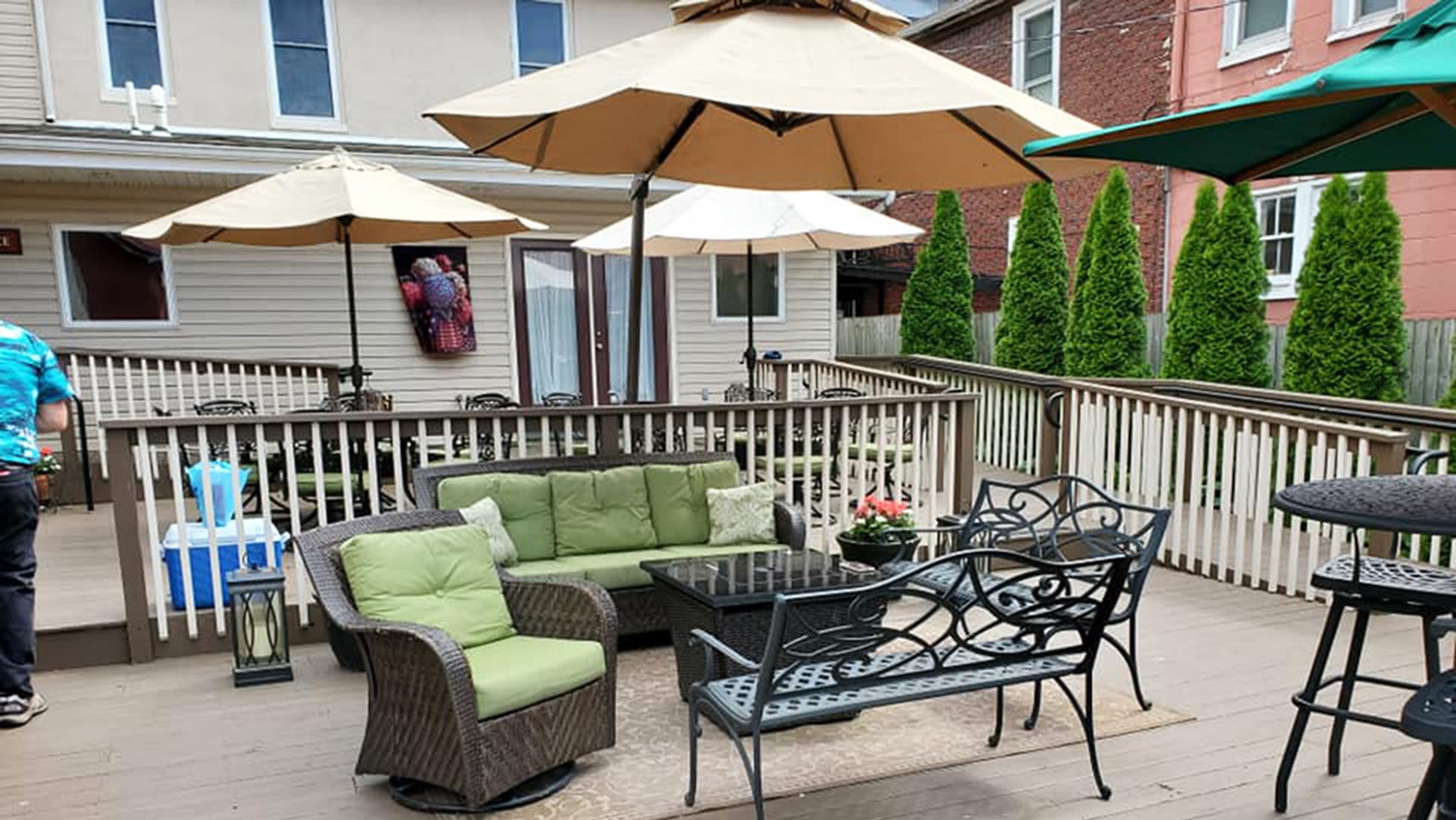 A cozy outdoor seating area with green cushions, tables, umbrellas, and surrounding greenery.