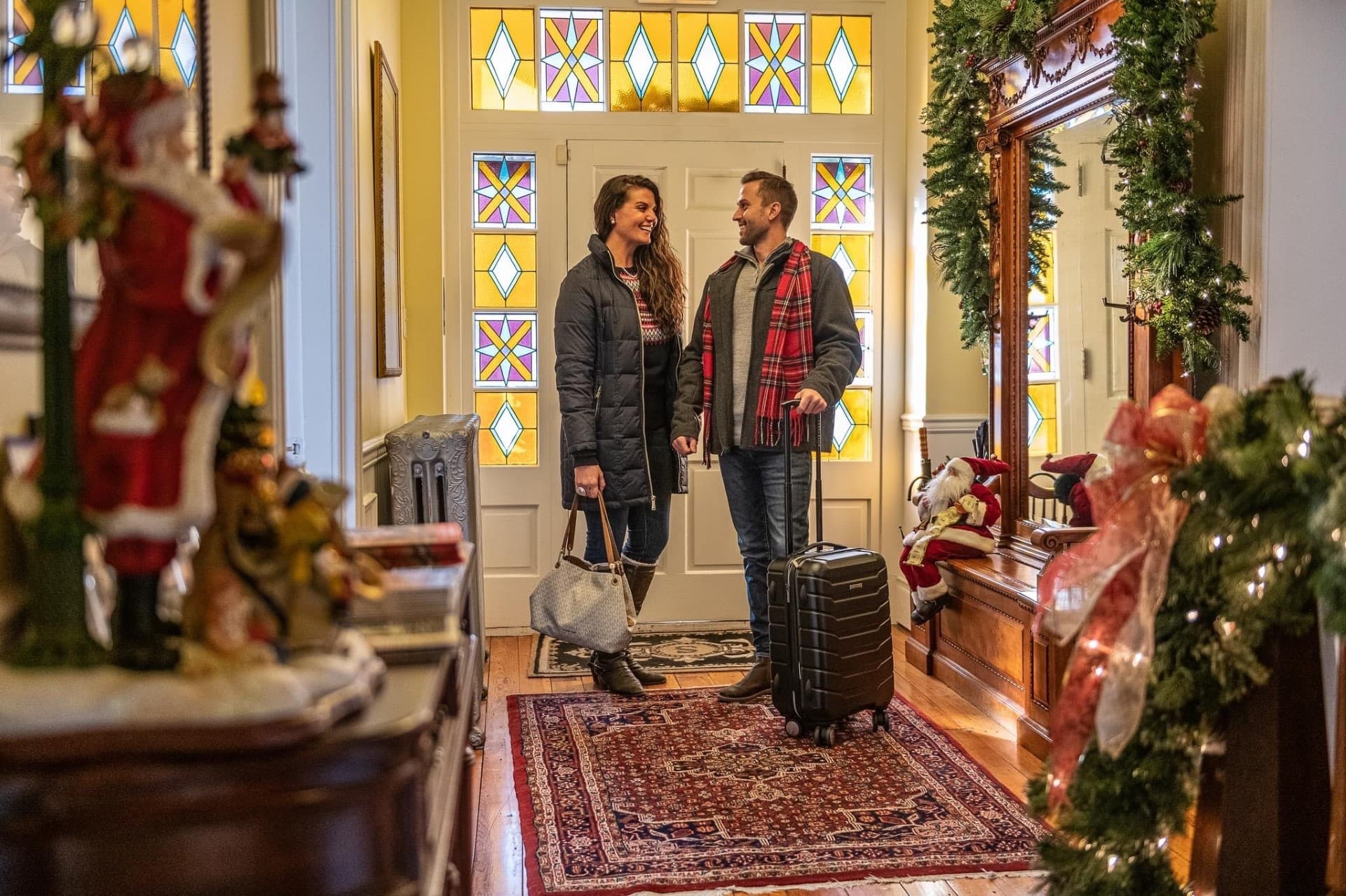 A couple stands together in a warmly decorated hallway, smiling and holding hands, with luggage nearby and holiday decorations around.