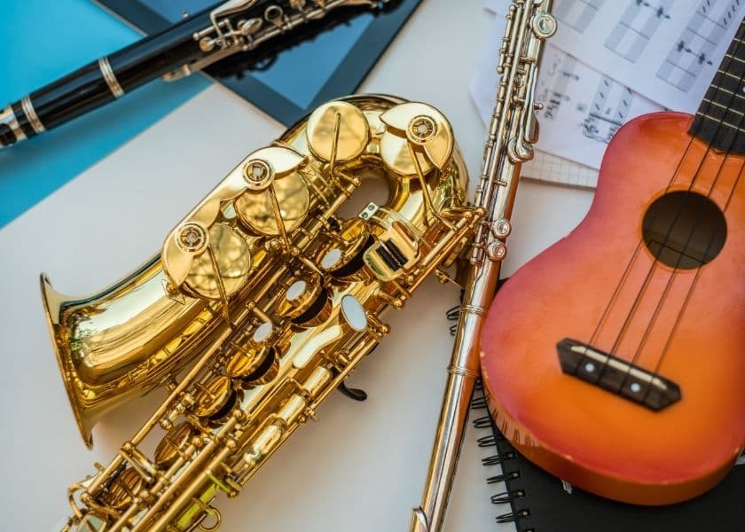 A collection of musical instruments including a saxophone, flute, and ukulele beside sheet music and a notebook. A collection of musical instruments including a saxophone, flute, and ukulele beside sheet music and a notebook.