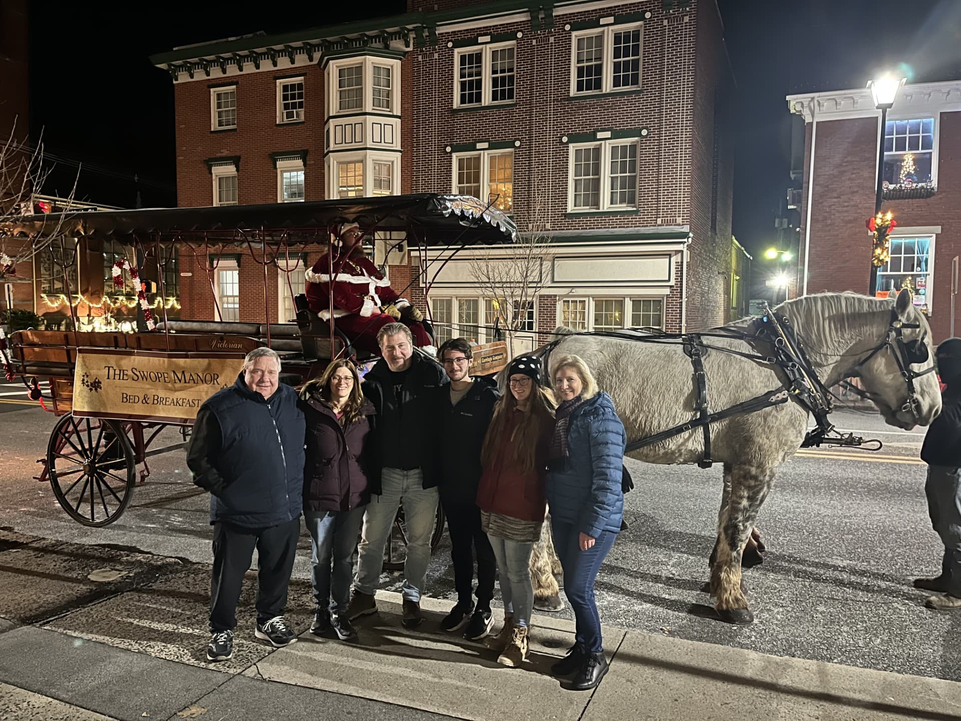 A group of six people poses with a horse-drawn carriage at night, featuring festive decorations and the Swope Manor sign.