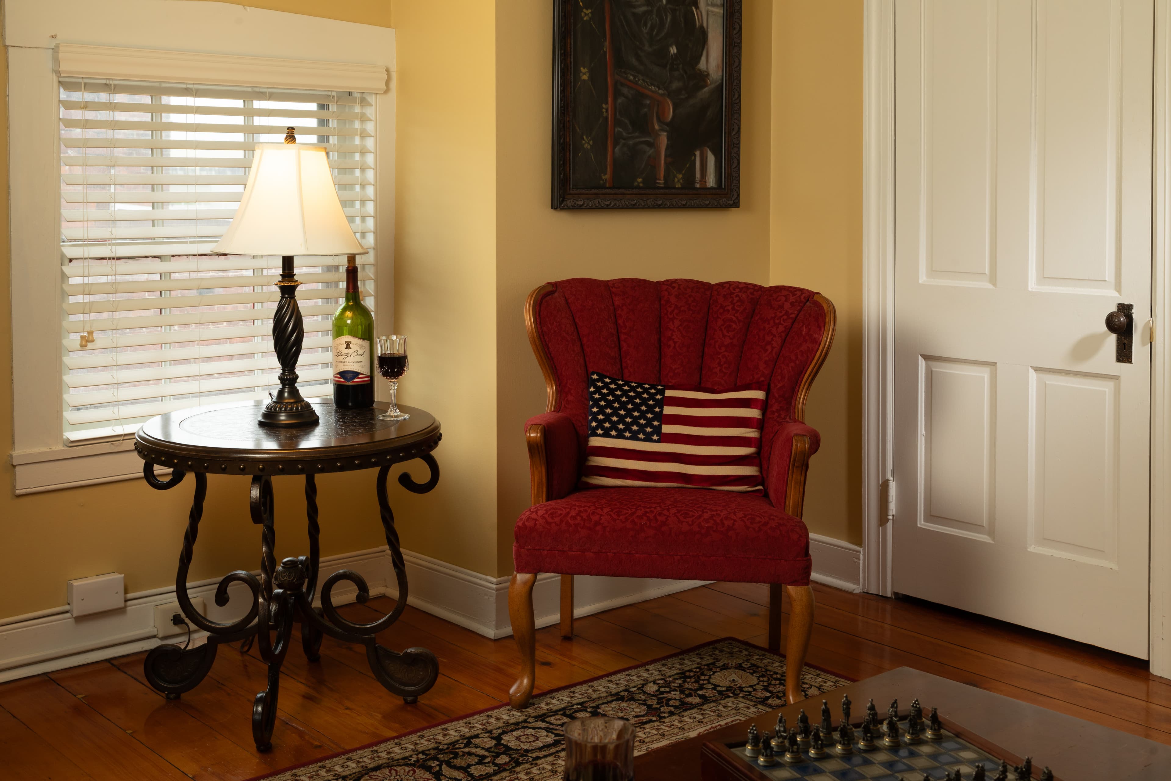 A cozy room featuring a red upholstered chair with an American flag pillow, a side table with a lamp and wine bottle, and a chess set on a patterned rug.