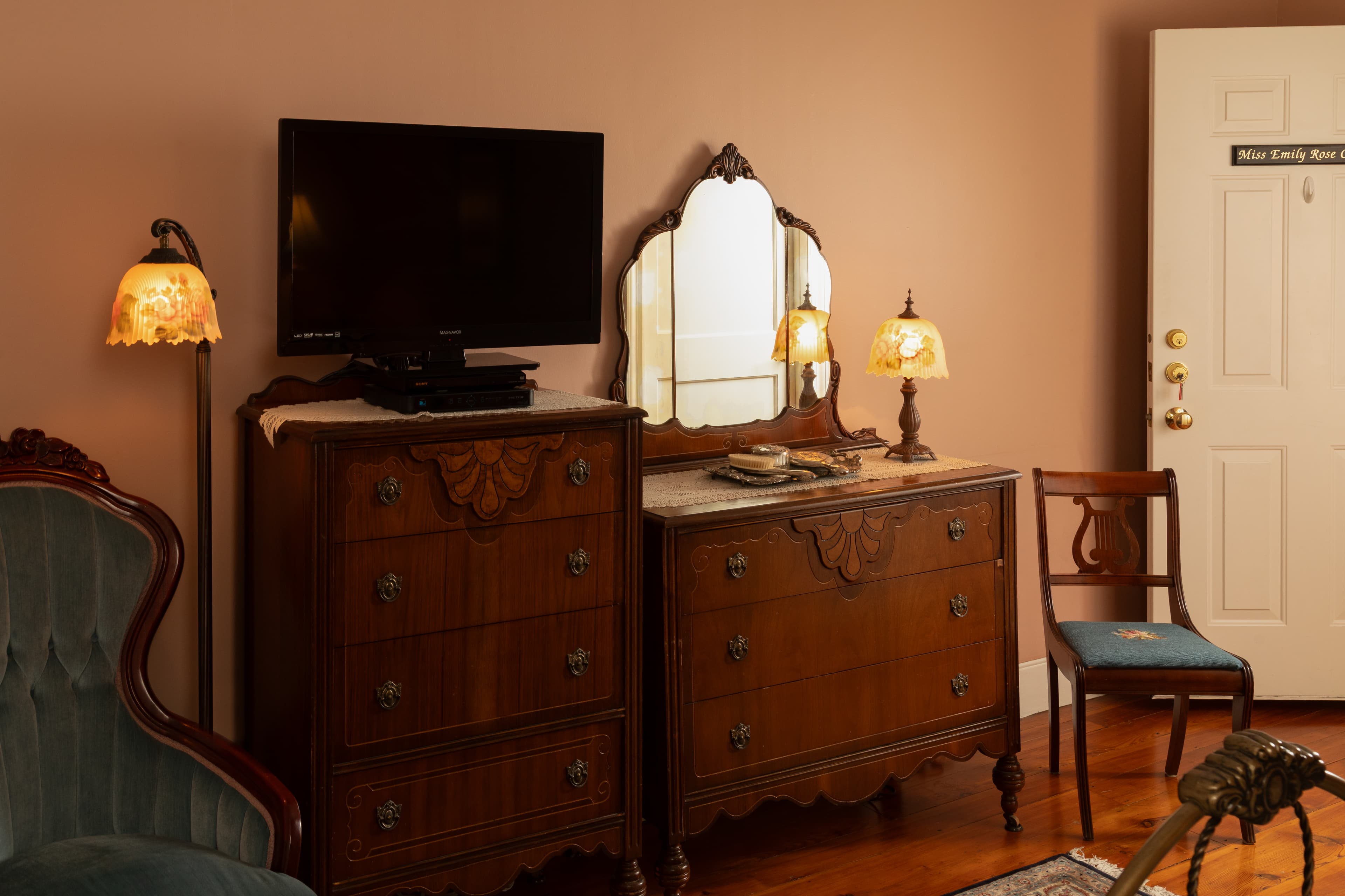 A vintage-style room features a dresser with a mirror, illuminated by two lamps, alongside a television and a chair.
