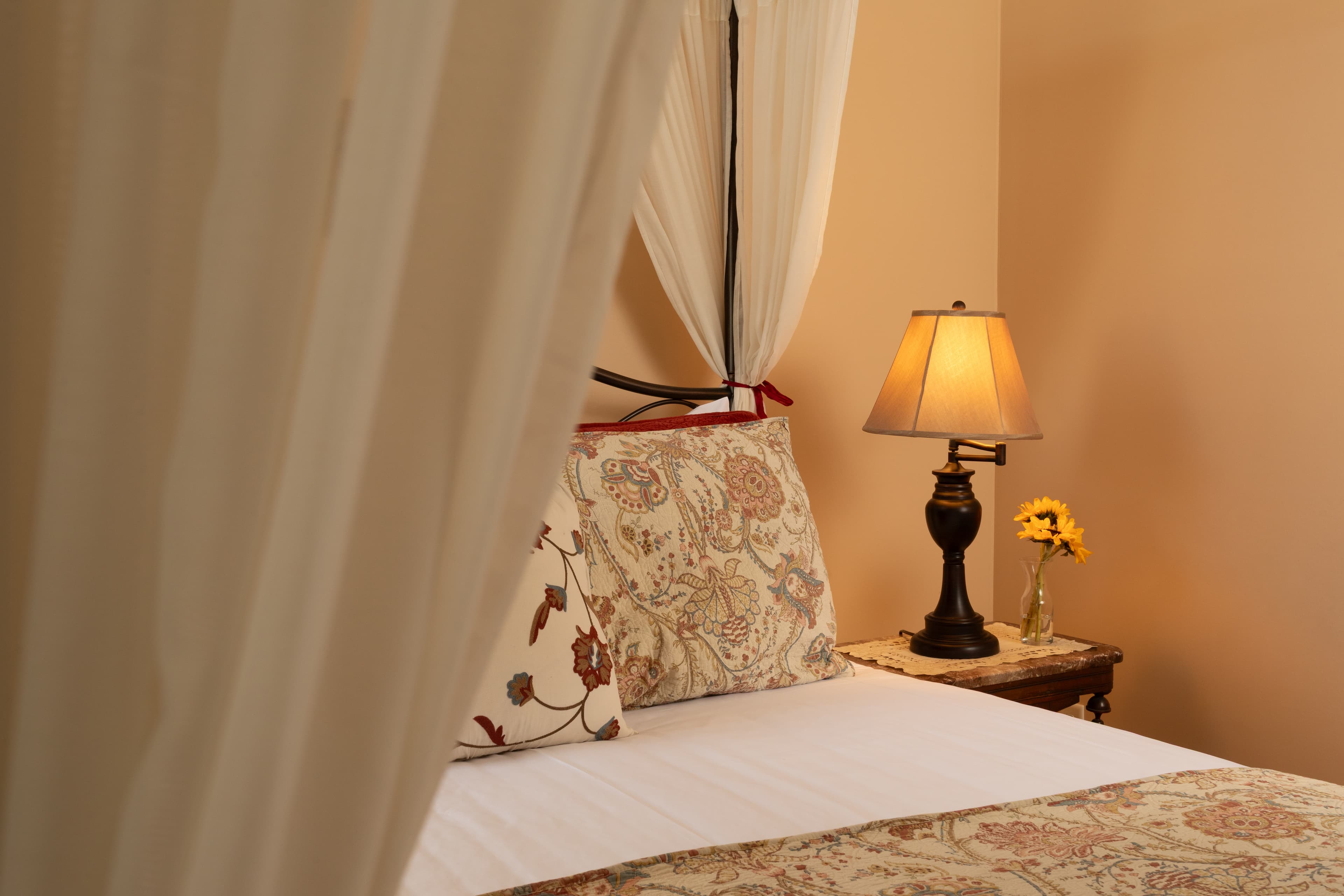 A cozy bedroom with a patterned bedspread, a lamp, and a vase of sunflowers.