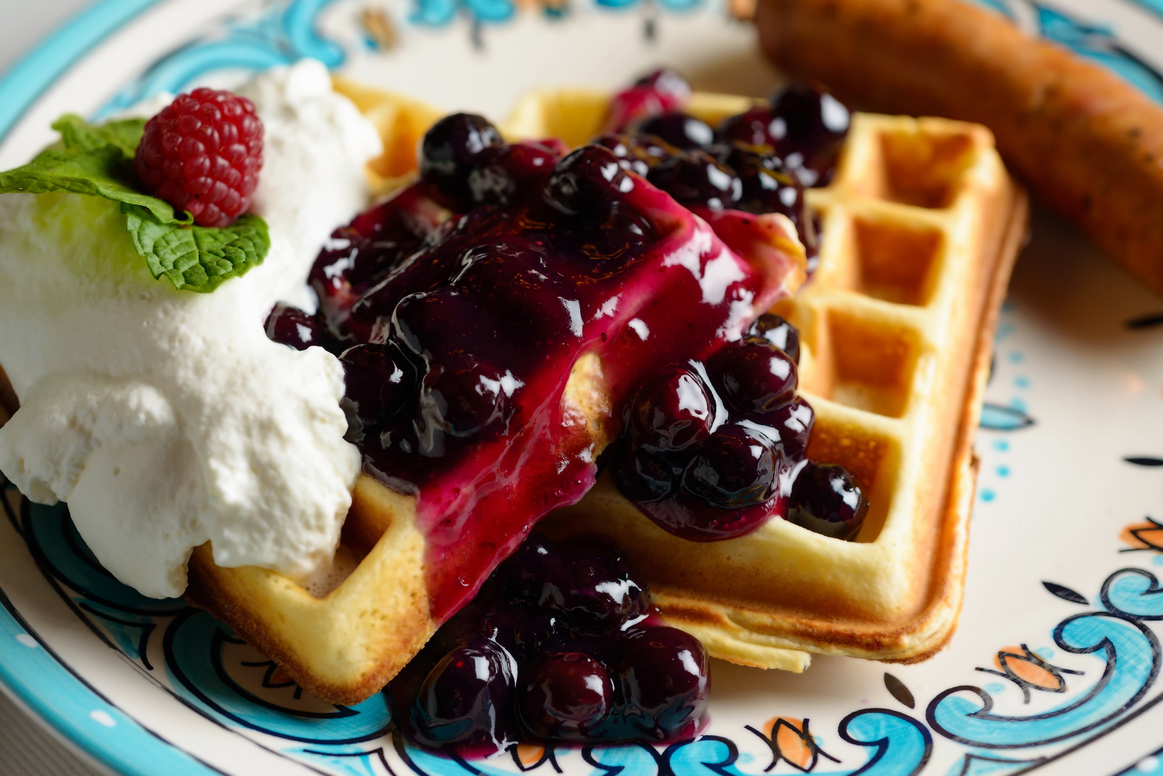 A waffle topped with blueberry sauce, whipped cream, and a raspberry, alongside a sausage link on a decorative plate.