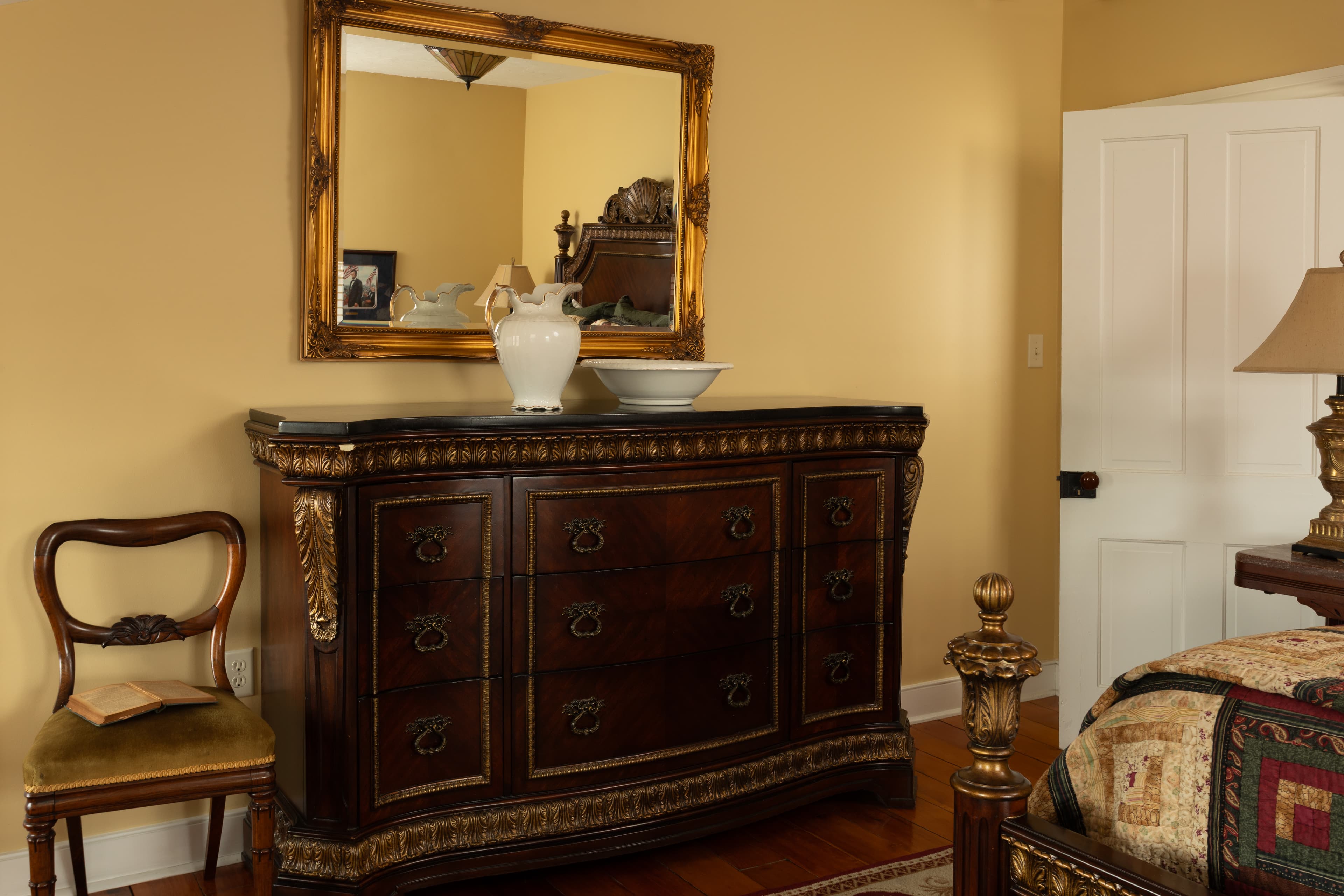A wooden dresser with decorative details, a mirror above, and a vase sits in a warmly colored bedroom.