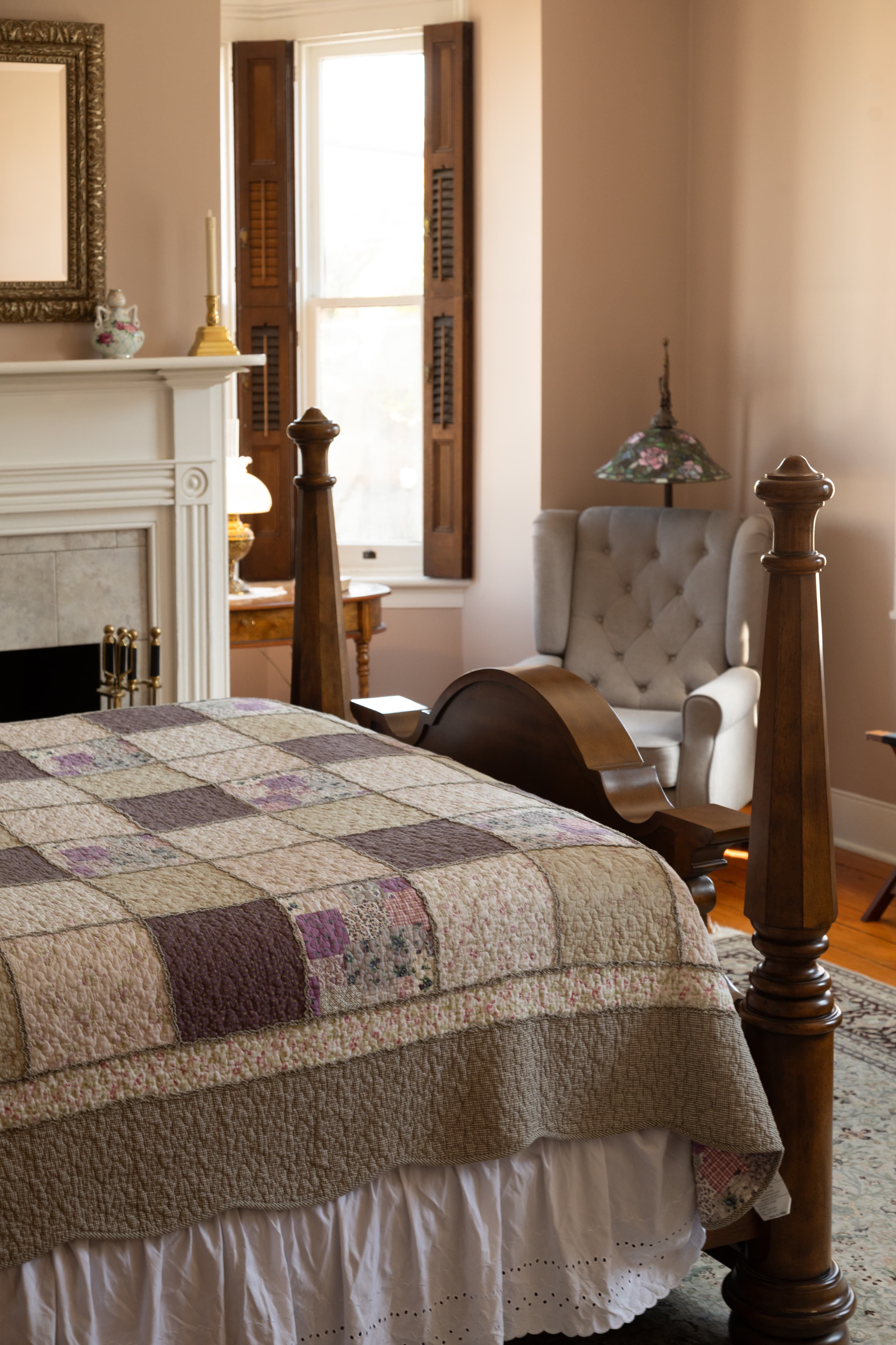 A cozy bedroom featuring a quilted bedspread, vintage armchair, and ornate fireplace.