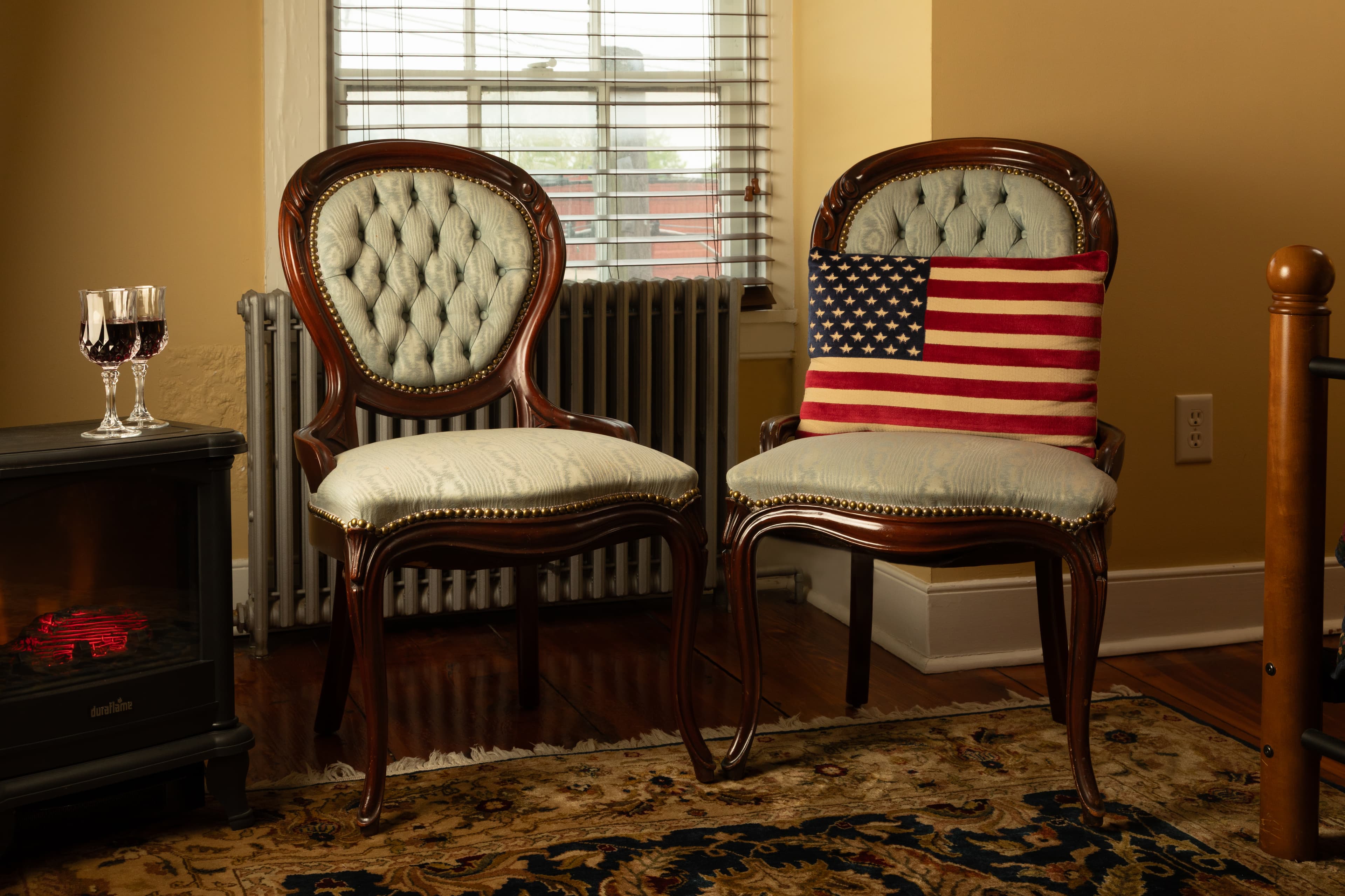 A cozy corner features two elegant chairs, one adorned with a pillow displaying the American flag, next to a small table with wine glasses and a decorative heater.