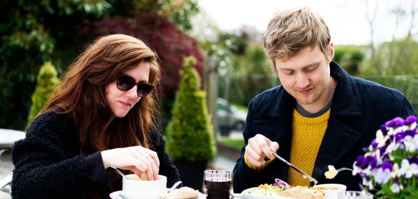 A couple wearing light jackets is dining al fresco with a spring bouquet of purple flowers on the table and lush, green foliage in the background.